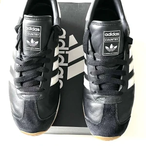 Adidas Country svarta sneakers - Klassiska Adidas Country sneakers i svart skinn med vita detaljer och de ikoniska tre ränderna på sidorna. Skorna har snörning, gummisula i naturfärg och rund tå. Perfekta för dig som gillar retrostil och sportig vibe.Klassiska Adidas Country OG sneakers i svart med vita ränder. En tidlös modell som passar till många olika stilar. Bekväma och stilrena. -Använd fåtal gånger är näst intill nyskick. -Adidas Skolådan medföljer. Storlek:40-40,5Passar Män och kvinnor. Ordpris:1397kr(Se sista bilden)
