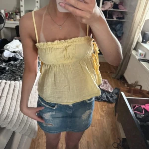 Gul linnetopp med knytband - Supersöt ljusgul linnetopp med smala axelband och volangkant upptill. Toppen har en luftig passform och knytband i sidan som ger en extra detalj. Perfekt för varma dagar och ger en fräsch sommarkänsla.