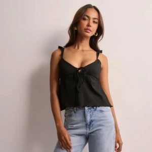 Nelly Svart linnetopp med knyt och volang  - Svart linnetopp med smala axelband och söt volangdetalj på axlarna. Toppen har v-ringning och knyt framtill, vilket ger en trendig look. Materialet är lätt och luftigt, perfekt för varma dagar. Passformen är normal och toppen är ärmlös. Den är helt oanvänd och är från Nelly. 