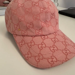 Rosa keps med GG-mönster - Säljer en snygg rosa keps med ikoniskt GG-mönster över hela kepsen. Justerbar rem baktill i matchande rosa och liten guldfärgad GG-detalj framtill. Kepsen är tillverkad i bomullstyg och har en klassisk böjd skärm.