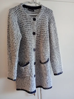 Grå fluffig cardigan från Busnel - Mysig och lång cardiganjacka från Busnel i grått fluffigt material. Jackan har rund halsringning, stora svarta knappar och två fickor framtill med mörka kanter. Perfekt för lager på lager och en avslappnad stil.