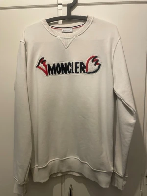 Vit sweatshirt från Moncler - Säljer en vit sweatshirt från Moncler med broderad logga i svart, rött och vitt på bröstet. Tröjan har rund halsringning, ribbade muddar och en avslappnad passform. Perfekt för dig som gillar streetwear och exklusiva märken. De står XL men den är väldigt liten i storleken passar bra på M