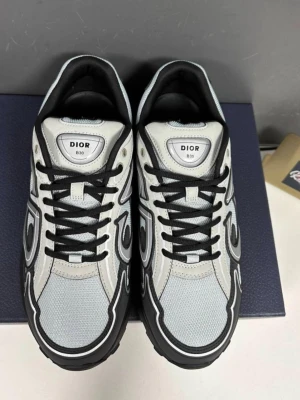Dior B30 sneakers i svart och vitt - Snygga Dior B30 sneakers i svart och vitt med coola grafiska detaljer och CD-logga på hälen. Ovandelen är i mesh och läder med svarta snören och robust sula. Perfekta för dig som gillar exklusiv streetwear och vill sticka ut med ett par riktigt trendiga skor. Pris kan diskuters (SKORNA ÄR LITE SMUTSIGARE ÄN DOM ÄR I BILDEN FAST KAN ENKELT RENSAS)