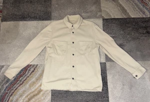 Beige overshirt från Arket storlek M - Stilren beige overshirt med två stora bröstfickor och svarta knappar framtill. Klassisk krage och långa ärmar med knapp vid manschetten. Perfekt lager-på-lager-plagg i mjukt tyg som ger en clean och modern look.
