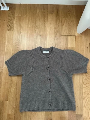 Grå stickad kofta från Wera - Kortärmad beige brun stickad kofta från Wera med rund halsringning och ribbade detaljer vid ärmslut, nederkant och hals. Koftan har fem knappar framtill och är gjord i ett mjukt, tjockt material som känns riktigt cozy.