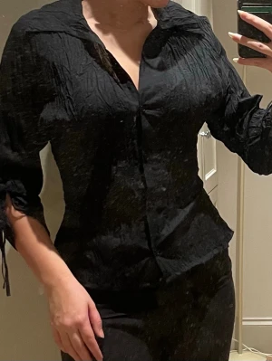 Svart crinkle-blus med knytärmar - Snygg svart blus med crinkle-effekt och figurnära passform. Blusen har långa ärmar med knytband vid ärmsluten och en djup v-ringning framtill. Perfekt för dig som gillar en stilren men ändå trendig look.