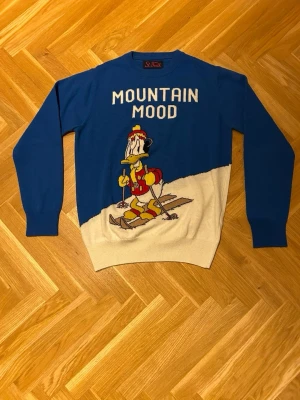 MC2 Saint Barth tröja - Blå och beige stickad MC2 Saint Barth tröja med texten 'MOUNTAIN MOOD' och en stor Kalle Anka-figur på skidor framtill. Tröjan har rund hals, långa ärmar och ribbade muddar. Perfekt för dig som gillar färgglada plagg