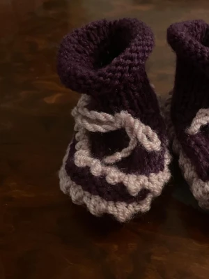 Stickade babytofflor i lila och rött - Två par handstickade babytofflor i mjukt garn. Ett par är lila med ljuslila detaljer och snören, det andra paret är ljuslila med röda och vita ränder samt snören. Perfekta för att hålla små fötter varma och mysiga. Säljs tillsammans.