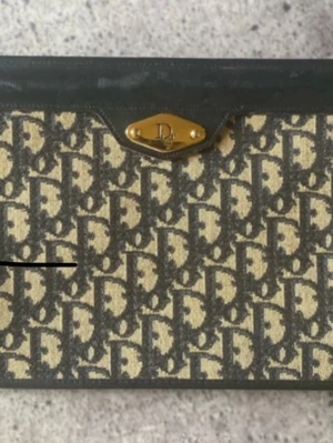 Dior portfölj i monogram canvas - Säljer en klassisk portfölj från Dior i beige och mörkbrun monogram canvas med detaljer i svart läder. Väskan har guldfärgad metallbricka med Dior-logga framtill och ikoniskt mönster över hela ytan. Perfekt för dig som gillar vintage och designer. B: ca 19 cm L: 19 cm