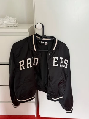 Svart Raiders bomberjacka - Snygg svart bomberjacka från H&M med vita detaljer och stora bokstäver 'RAIDERS' på framsidan. Jackan har ribbade muddar vid ärmslut, krage och nederkant samt tryckknappar framtill. Perfekt för en sportig och trendig look.