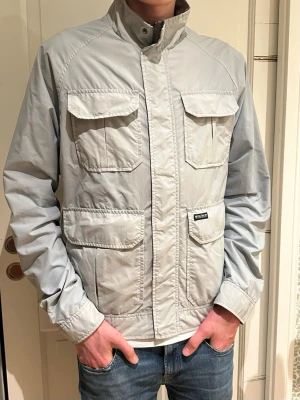 Ljusgrå vindjacka från woolrich  - Snygg ljusgrå vindjacka från woolrich med fyra stora fickor framtill och hög krage. Jackan har dragkedja och tryckknappar, samt justerbar nederkant. Perfekt för dig som gillar stilren och funktionell design.
