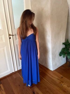 Blå axelbandslös långklänning - En stilren blå axelbandslös långklänning med v-ringad överdel och lätt rynkad kjol. Klänningen har en flowy passform och är perfekt för sommarens alla tillfällen. Tillverkad i ett mjukt och följsamt material som ger en elegant siluett.
