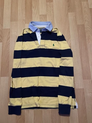 Randig tröja från Ralph Lauren - Säljer en klassisk långärmad tröja från Ralph Lauren med breda marinblå och gula ränder. Tröjan har vit knappslå, ljusblå krage och den ikoniska gröna polospelaren broderad på bröstet. Tillverkad i mjuk bomull för en avslappnad men stilren look.