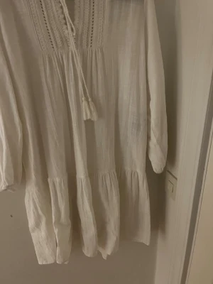 Vit klänning med broderingar - Luftig vit klänning i boho-stil med broderade detaljer vid halsen och knytband med tofsar. Klänningen har långa ärmar och volang nedtill, tillverkad i ett tunt, vävt material som ger en avslappnad känsla.