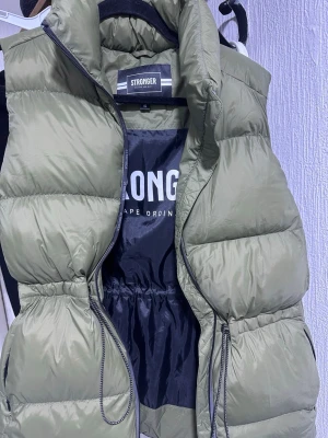 Olivgrön dunväst från Stronger - Snygg olivgrön sopuffad dunväst från Stronger med hög krage och dragkedja framtill. Västen har justerbar midja med snörning och två fickor med dragkedja. Perfekt för lager på lager och streetstyle.