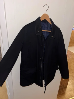 Svart rock från Rose & Born - Stilren svart rock från Rose & Born med klassisk krage och flera fickor framtill. Rocken har en quiltad insida för extra värme och stängs med dragkedja och knappar. Perfekt för dig som vill ha en tidlös och snygg look under kallare dagar. Nypris 23 000 kr