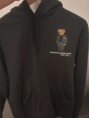 Svart hoodie från Polo Ralph Lauren - Svart hoodie från Polo Ralph Lauren med den klassiska björnen broderad på bröstet. Hoodien har huva med dragsko och en stor magficka framtill. Tillverkad i mjukt bomullsmaterial som är skönt att bära. Perfekt för en avslappnad och stilren look.