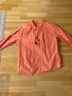 Korallfärgad skjorta med knappar - Säljer en snygg korallfärgad skjorta med lång ärm och klassisk krage. Skjortan har knäppning framtill och en bröstficka. Perfekt för dig som vill ha en färgklick i garderoben!
