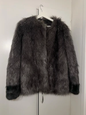 Mörkgrå pälsjacka med svarta detaljer - Snygg mörkgrå pälsjacka i fuskpäls med svarta detaljer vid ärmsluten. Jackan har rak passform, lång ärm och stängs framtill. Perfekt för att hålla värmen under kalla dagar och ger en lyxig känsla till din outfit.