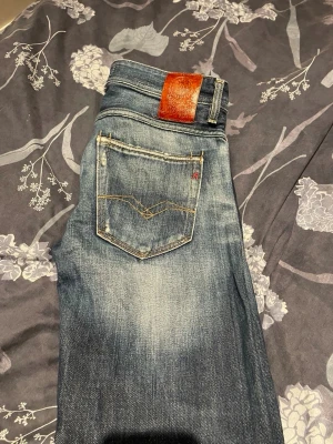 Blå slitna jeans från Replay - Säljer ett par blå jeans från Replay med snyggt slitna detaljer och klassisk femficksmodell. Jeansen har en skön tvättad look, kontrastsömmar och ett brunt läderpatch bak i midjan. Perfekta för en avslappnad och trendig stil.