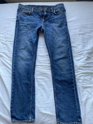 Blå  jeans från Nudie Jeans - Säljer ett par klassiska blå jeans från Nudie Jeans i modellen Lean Dean. Jeansen har snygga slitningar och kontrastsömmar samt Nudie-logga på baksidan. Perfekta för en avslappnad stil. Storlek W32 L32