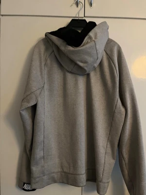 Grå hoodie från Nike med dragkedja - Snygg grå hoodie från Nike med svart innerfoder i huvan och dragkedja framtill. Klassisk design med Nike-logga på insidan av kragen och mjukt material som passar perfekt till vardagslooken. Huvan ger extra streetkänsla och komfort.