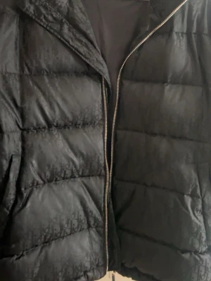 Svart dunjacka från Dior - Snygg svart dunjacka från Dior med diskret monogrammönster över hela jackan. Jackan har en rak passform, dragkedja framtill och är tillverkad i ett glansigt material som ger en exklusiv känsla. Perfekt för kalla vinterdagar och för dig som vill ha en lyxig look. Inget kvitto, 