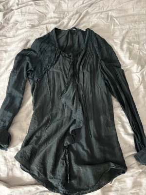 Mörk blå/grön blus med spetsdetaljer - Snygg mörk blå/grön blus med långa ärmar och spetsdetaljer vid axlarna. Blusen har en lös passform, volangkant framtill, knappar fram och knytband vid halsen. Tillverkad i ett tunt, lätt material som ger ett luftigt intryck.