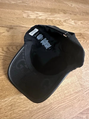 Svart keps från Gucci med logga - Snygg svart keps från Gucci med klassisk böjd skärm och diskret GG-mönster på ovansidan. Justerbar rem bak i svart läder med präglad Gucci-logga. Tillverkad i en mix av textil och läder för en exklusiv känsla.
