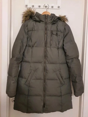 Lång grå dunjacka med fuskpäls - Snygg lång grå dunjacka med huva och fuskpälsdetalj. Jackan har dragkedja framtill, bröstficka med dragkedja och två sidofickor. Quiltad design som håller värmen och ger en modern look. Perfekt för kalla vinterdagar.