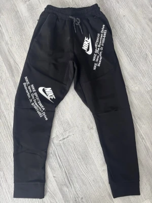 Svarta Nike tech-fleece byxor med tryck - Säljer ett par svarta tech-fleece byxor från Nike med vita loggor och texttryck på både höger och vänster ben. Byxorna har resår i midjan med snörning och muddar vid bensluten. Perfekta för chill dagar eller träning. Materialet känns mjukt och bekvämt. Storlek M men passar XS/S