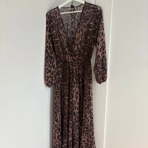 Lång leopardmönstrad omlottklänning - Säljer en långklänning med leopardmönster och omlottdesign. Klänningen har v-ringning, långa ärmar och resår i midjan för en snygg passform. Materialet är tunt och flowy, perfekt för dig som vill sticka ut.