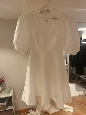 Vit studentklänning med puffärm - Superfin vit kort klänning med puffärmar och v-ringning. Klänningen har en lätt glansig yta och är tillverkad i ett satinliknande material som ger en lyxig känsla. Perfekt för dig som tar studenten iår