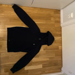 Polo Hoodie  - Klassisk svart hoodie med dragkedja framtill och huva. Hoodien har långa ärmar, ribbade muddar och fickor på sidorna. Perfekt för en avslappnad och stilren look. Passar till det mesta och är enkel att matcha.