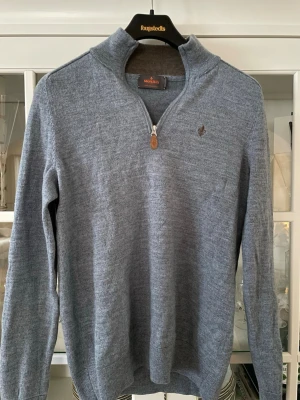 Blå halvzip tröja från Morris - OBS! Liten i modellen (XS är mer korrekt). Snygg blå stickad tröja från Morris i 100% merinoull. Tröjan har halv dragkedja, ribbade muddar och diskret logga broderad på bröstet. Perfekt för lager på lager och ger en clean look.