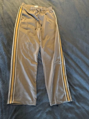 Just Junkies byxor - Säljer ett par beige Just Junkies mjukisbyxor med svarta och vita sidoremsor. Byxorna har resår i midjan med dragsko och raka ben. Materialet är mjukt och följsamt, perfekt för chill dagar. En bakficka med dragkedja finns också.