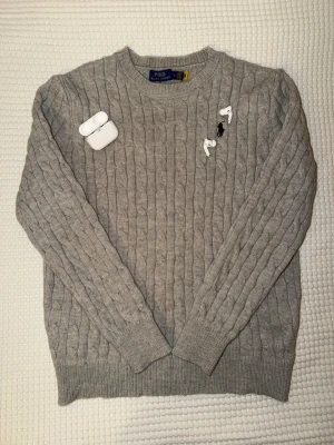 Ralph Lauren Sweater - Kabelstickad grå tröja från Polo Ralph Lauren med klassisk rund halsringning och svart broderad logga på bröstet. Tröjan har ribbade muddar vid ärmslut och nederkant, och är tillverkad i mjukt material som passar perfekt till höst och vinter.
