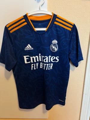 Real Madrid bortatröja blå Adidas - Snygg blå Real Madrid bortatröja från Adidas med gula detaljer på axlarna och klubbmärke på bröstet. Tröjan har korta ärmar, V-ringning och tryckta loggor samt sponsortexten 'Emirates Fly Better' på framsidan. Tillverkad i lätt och funktionellt material. Storlek 158/164 13-14 år