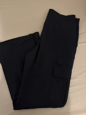 Svarta cargobyxor med fickor - Säljer ett par svarta cargobyxor med klassiska sidofickor och bälteshällor. Byxorna har en rak passform och är tillverkade i ett mjukt, stretchigt material som känns bekvämt hela dagen. Perfekta för dig som gillar en clean och stilren look.