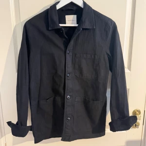Svart overshirt från A Day's March - Stilren svart overshirt från A Day's March i kraftigt bomullstyg. Klassisk krage, tre framfickor och knäppning med svarta knappar. Perfekt lager-på-lager-plagg med avslappnad passform och uppvikta ärmslut.