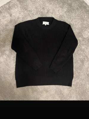 Svart stickad tröja Maison Margiela - Säljer en svart stickad tröja från Maison Margiela. Tröjan har rund halsringning, ribbstickad struktur och långa ärmar. Perfekt för dig som gillar minimalistisk och stilren design. Materialet känns mjukt och håller värmen bra.