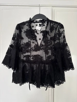 Svart transparent spetsblus H&M - Snygg svart blus i transparent spets från H&M med blommigt mönster. Blusen har v-ringning, volangkant nedtill och vida trekvartsärmar. Perfekt för att skapa en trendig lager-på-lager-look. Materialet är lätt och luftigt.