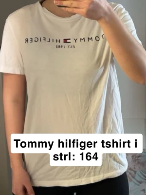 Vit Tommy Hilfiger t-shirt - Klassisk vit t-shirt från Tommy Hilfiger med logga och text tryckt över bröstet. T-shirten har rund halsringning och korta ärmar. Tillverkad i mjuk bomull tyvärr skrynklig då den inte används av mig men går att stryka eller tvätta så e problemet löst😊