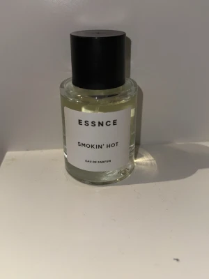 ESSNCE Smokin' Hot Eau de Parfum - ESSNCE Smokin' Hot Eau de Parfum kommer i en stilren, rund glasflaska med svart lock och vit etikett. Doften är elegant förpackad och flaskan har en minimalistisk design som passar perfekt på parfymhyllan.
