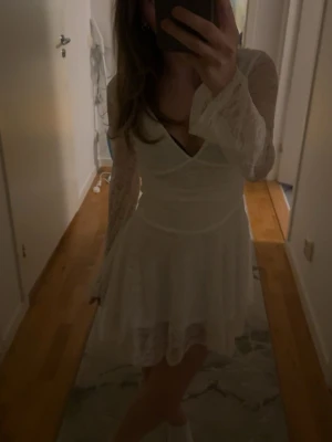 Vit studentklänning med öppen rygg - Säljer en superfin vit kortklänning i spets med långärmade ärmar och v-ringad front. Klänningen har en snygg öppen rygg och volang i kjolen, vilket ger en romantisk vibe. Perfekt för sommarens alla tillfällen! Vara testad från dressy