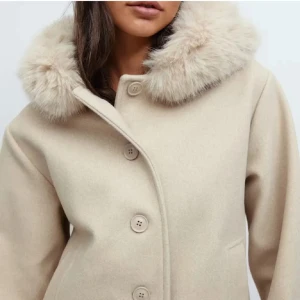 Beige kappa med fuskpäls - Stilren beige kappa med stor, fluffig fuskpäls runt huvan och knäppning framtill. Kappan har en rak passform och diskreta fickor i sidorna. Perfekt för dig som vill ha en trendig och mysig look under kalla dagar.