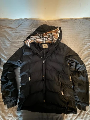 Svart pufferjacka från Burberry - Snygg svart pufferjacka från Burberry med klassiskt rutigt foder i huvan. Jackan har dragkedja, knappar, två framfickor med dragkedja och ribbade muddar vid ärmsluten. Perfekt för kalla vinterdagar och riktigt stilren.