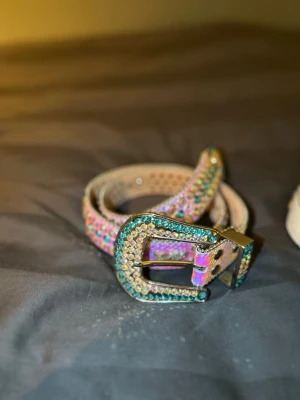 Tuttolente bälte - Säljer ett smalt bälte täckt av glittriga, färgglada stenar och med ett stort, dekorerat spänne. Bältet har en unik och iögonfallande design som verkligen sticker ut. Perfekt för dig som vill addera lite extra bling till din outfit.