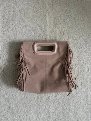 Beige kuvertväska med fransar - Säljer en beige kuvertväska i mockaimitation med dekorativa fransar på sidorna och ett rektangulärt handtag upptill. Väskan har en mjuk och stilren look som passar perfekt till din outfit. Den är från maje och ordinarie pris är 2800kr 