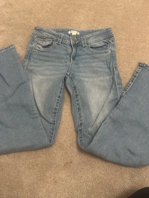 Ljusblå bootcut jeans - Säljer ett par ljusblå bootcut jeans med klassisk femficksdesign och slitningar framtill. Jeansen har normal midja och dragkedja med knapp. Perfekta för en avslappnad och trendig look.